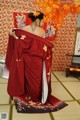 A woman in a red kimono standing in front of a wall of flowers.