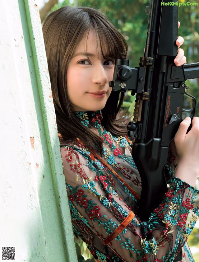 A woman holding a rifle leaning against a wall.